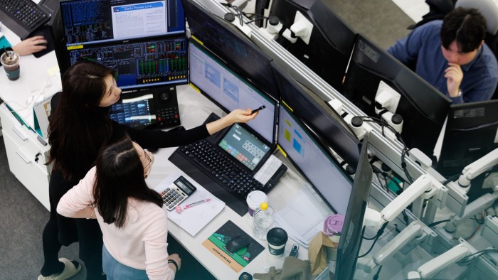 Employees in a trading room at Hana Bank in Seoul work at desks with multiple computer monitors displaying financial data. One person points at a screen with a pen, while another observes closely