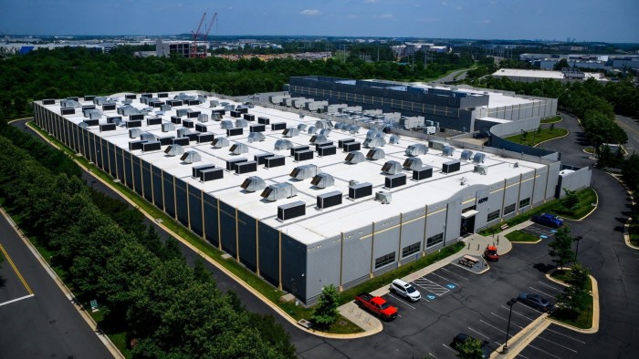 Aerial view of a large data center facility in Sterling, Virginia, surrounded by greenery and parking lots. The building features numerous HVAC units on the roof, indicating high energy usage for cooling.