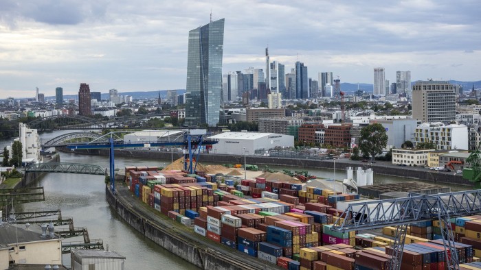 A container ship sits at a port in Frankfurt. The skyline is visible in the background