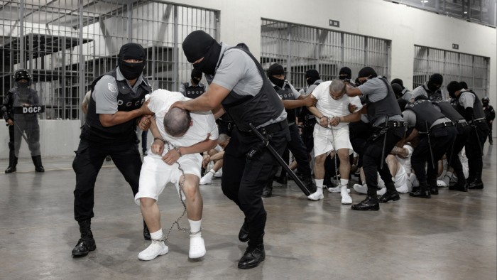 Salvadoran police officers, wearing black masks and uniforms marked "DGCP," escort several handcuffed individuals in white prison attire through a detention facility.